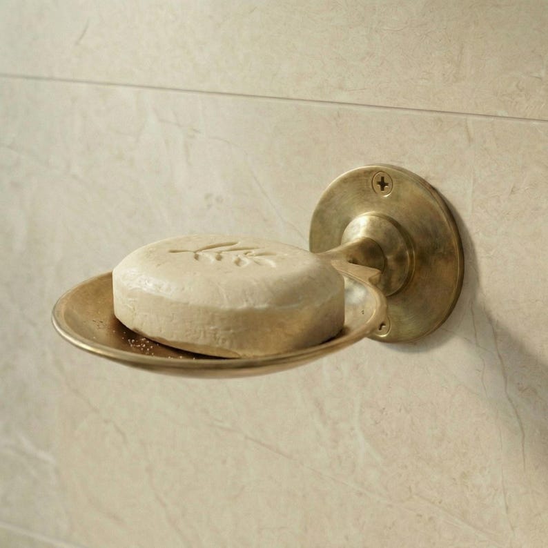 May include: A brass soap dish with a round, wall-mounted backplate and a curved tray. A round, light-colored bar of soap with an embossed design rests in the dish. The fixture is mounted on a beige tiled wall.