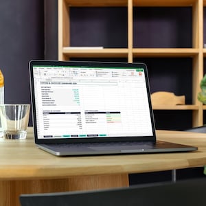 May include: A laptop displaying a spreadsheet sits on a wooden table, next to a glass of water with orange slices and an empty glass. The laptop screen shows data in a spreadsheet format. A wooden shelf and a plant are in the background.