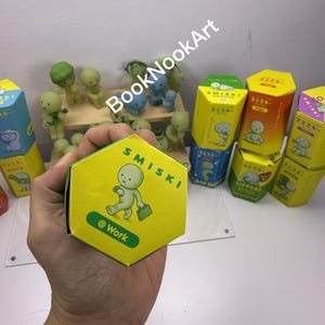A person's hand holding a yellow hexagonal box with a cartoon character on it, surrounded by various colorful boxes and figurines.