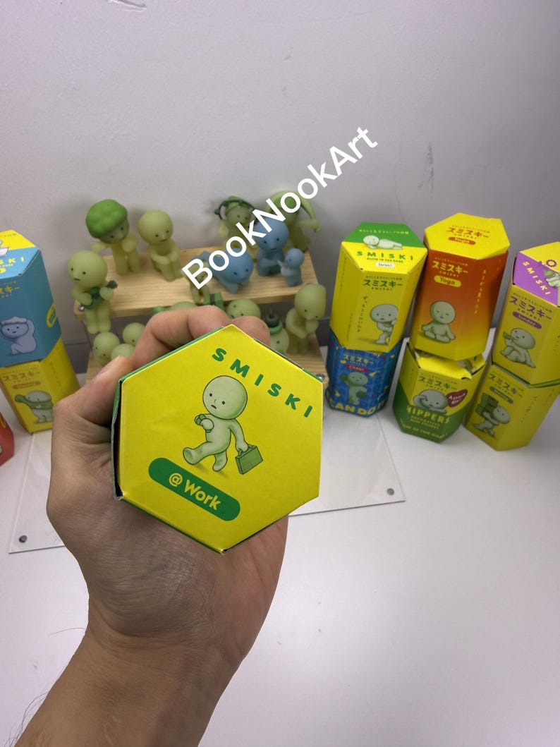 A person's hand holding a yellow hexagonal box with a cartoon character on it, surrounded by various colorful boxes and figurines.