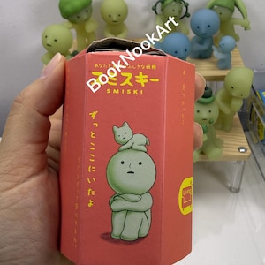 A hand holding a red box with a cartoon character on it, surrounded by various other small figurines.