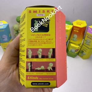 A hand holding a yellow box with a picture of a living toy, surrounded by other similar boxes.