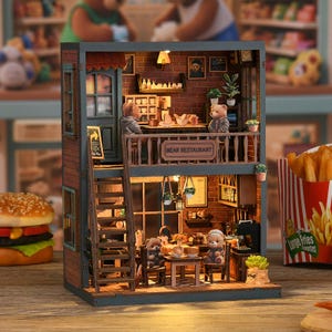 以下が含まれることがあります： 2階建てのミニチュア木製レストラン「Bear Restaurant」。バルコニーと梯子が付いています。内部には小さなテーブル、椅子、クマの置物が配置されています。レストランはレンガ造りの外観で、小さな植物や額縁で装飾されています。
