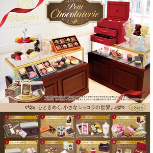 Op de afbeelding: Een miniatuurdisplay van chocolade en zoetwaren, met een verscheidenheid aan chocolade in dozen, gebak en snoep. De display bevat een rode kast met laden, glazen vitrines en een etagère. De tekst "Petit Chocolaterie" is zichtbaar.