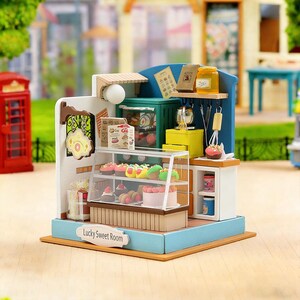 Könnte beinhalten: Miniatur-Bäckerei-Diorama mit blau-weißer Fassade, mit einer Vitrine voller bunter Backwaren, einem kleinen Ofen und Regalen mit Gläsern und Utensilien. Auf der Basis steht "Lucky Sweet Room."