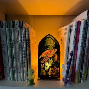 May include: A decorative stained-glass style lamp featuring a sea turtle design. The lamp is illuminated, showcasing the turtle and aquatic plants. It sits on a shelf with books and small figurines, creating a warm, inviting ambiance.