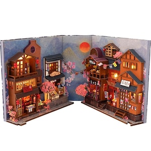May include: A miniature Japanese village diorama with detailed wooden buildings, illuminated windows, and cherry blossom accents. The scene includes a backdrop of a sunset and a night sky, creating a charming and detailed display.