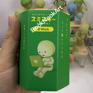 A hand holding a green box with a cartoon character on it, surrounded by various figurines.