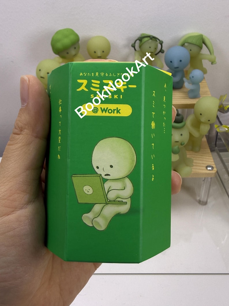 A hand holding a green box with a cartoon character on it, surrounded by various figurines.