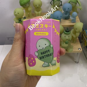 Op de afbeelding: Een hand houdt een roze zeshoekige doos met Japanse tekst en het woord "Birthday" in het geel. De doos heeft een cartoonfiguur met een groen hart met de tekst "Happy Birthday". Verschillende kleine, grillige beeldjes in verschillende kleuren staan op een plank op de achtergrond.