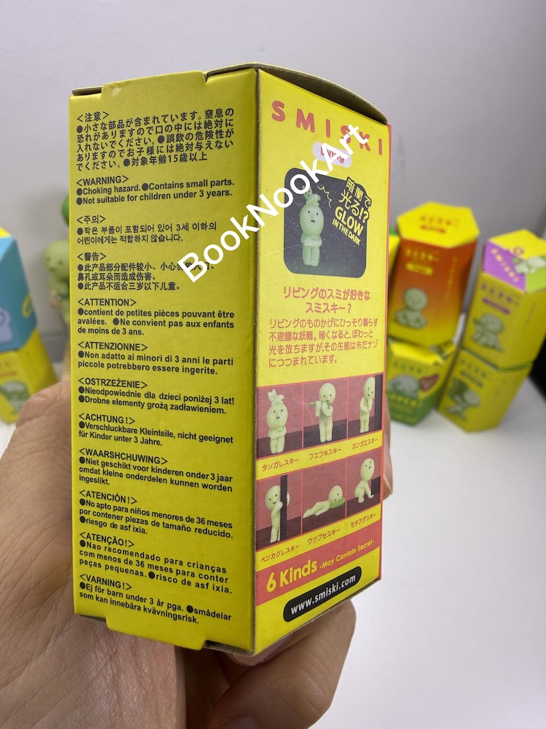 A hand holding a yellow box with text and images, including a small figurine and various safety warnings.