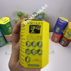 A hand holding a yellow box with Japanese text and images, surrounded by various colorful boxes and figurines.