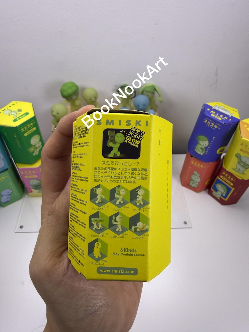 A hand holding a yellow box with Japanese text and images, surrounded by various colorful boxes and figurines.