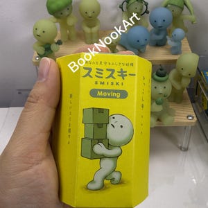 A hand holding a yellow box with a cartoon character on it, surrounded by various figurines.