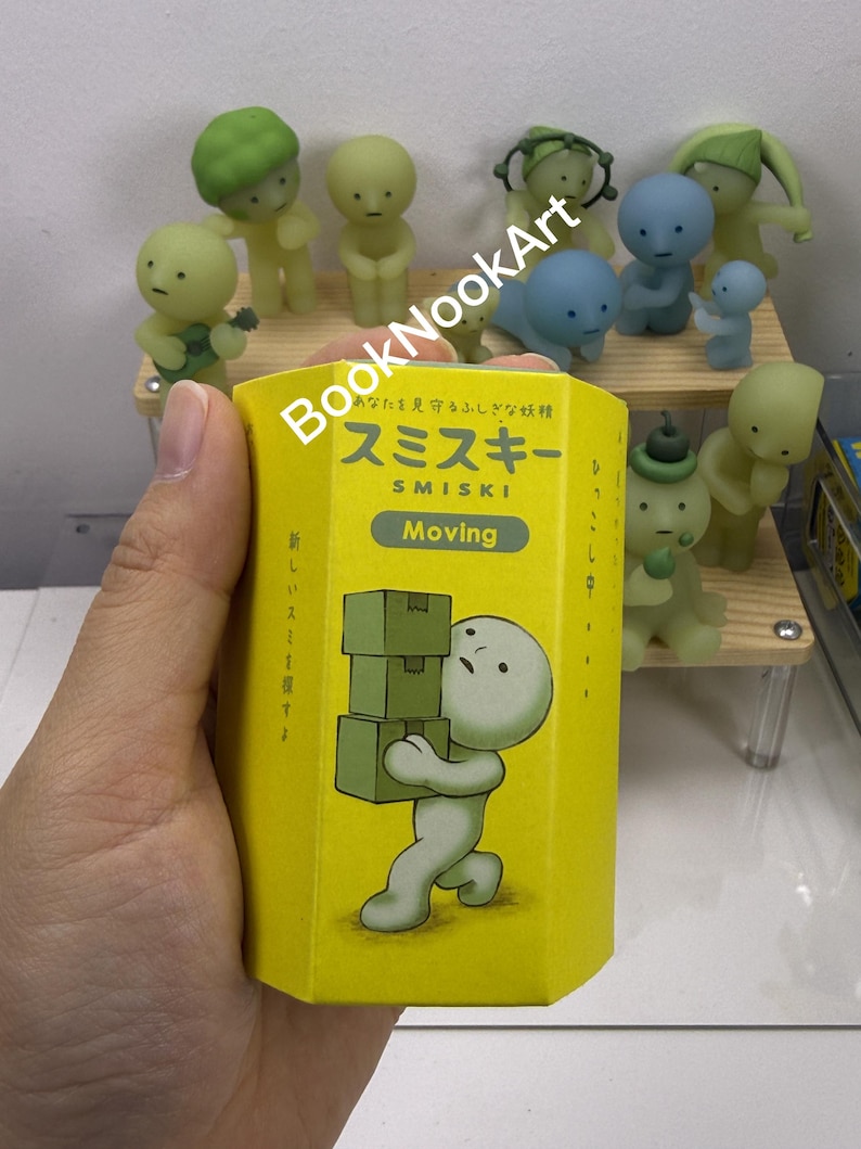 A hand holding a yellow box with a cartoon character on it, surrounded by various figurines.