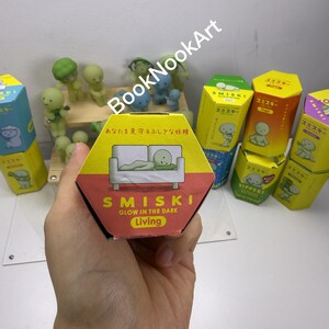 A hand holding a hexagonal box with a cartoon image of a green frog on it, surrounded by various colorful boxes and figurines.