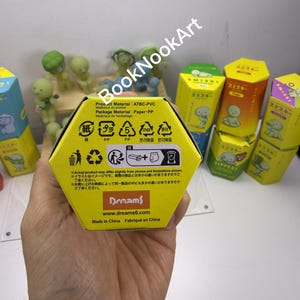 A hand holding a yellow box with various symbols and text, including recycling and disposal information, in front of a collection of colorful boxes and figurines.