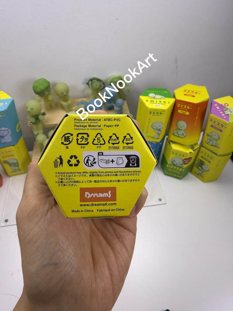 A hand holding a yellow box with various symbols and text, including recycling and disposal information, in front of a collection of colorful boxes and figurines.