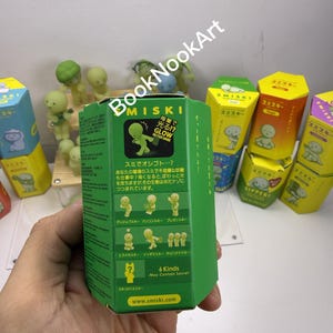 A person's hand holding a green box with Japanese text and illustrations, surrounded by various colorful boxes and figurines.