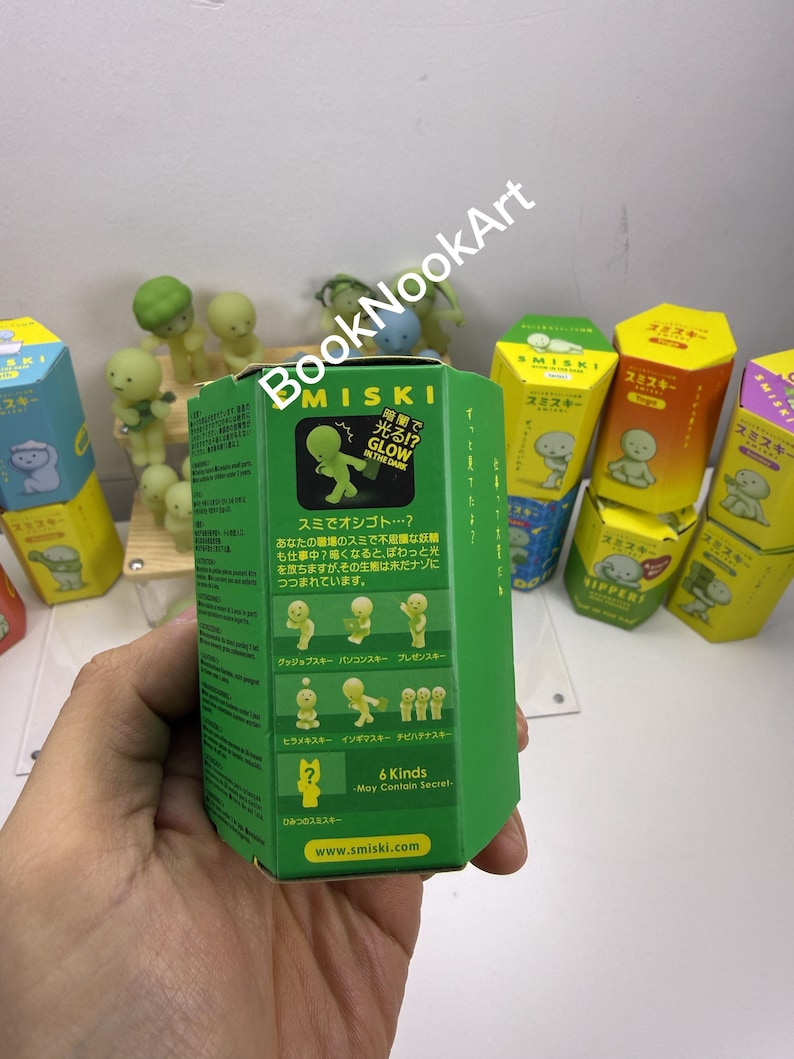 A person's hand holding a green box with Japanese text and illustrations, surrounded by various colorful boxes and figurines.