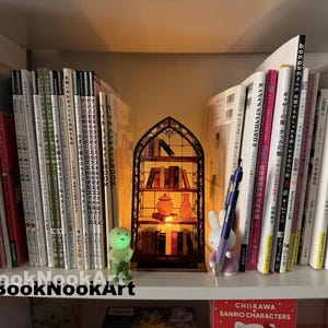 May include: A bookshelf filled with books and a decorative book nook. The book nook is a miniature, illuminated scene with a stained-glass window design, containing small books and a vase. A green figurine and a purple bunny figurine are also on the shelf.