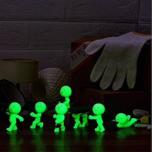 A group of small, glowing green figurines are arranged on a dark surface, with a pair of white gloves and a lamp visible in the background.