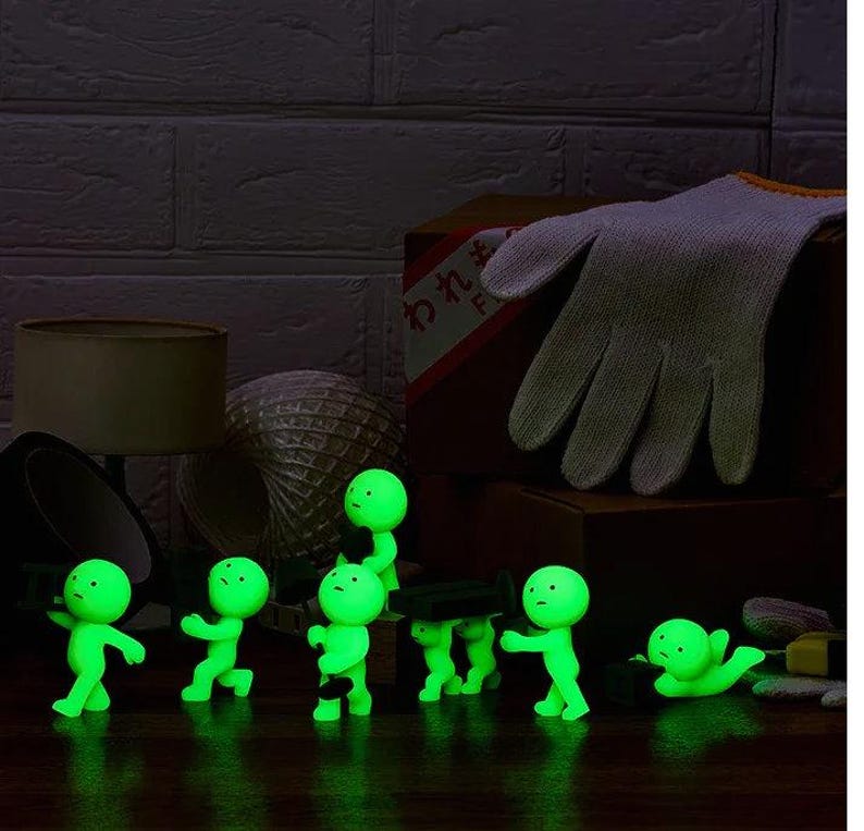 A group of small, glowing green figurines are arranged on a dark surface, with a pair of white gloves and a lamp visible in the background.