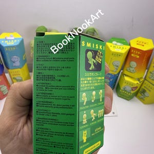 A person's hand holding a green box with text and illustrations, with colorful boxes in the background.