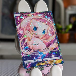 May include: A holographic Mew ex trading card featuring a pink character with blue eyes, displayed on a white and light green rabbit-shaped stand. The card shows the text Mew ex, 130 HP, Psyshot, and Genome Hacking against a colorful cosmic background.