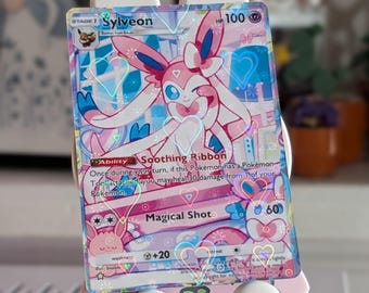 May include: A Sylveon trading card featuring a holographic finish with heart-shaped patterns. The card displays the character Sylveon, 100 HP, the Soothing Ribbon ability, and the Magical Shot attack. The design includes pink and blue colors.