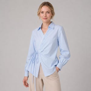 May include: A light blue long-sleeved button-down shirt featuring a wrap-style front with a side tie closure and delicate white lace trim along the diagonal placket. The shirt has a classic collar, cuffed sleeves, and is paired with beige trousers.