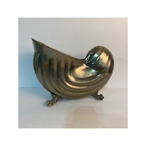 May include: A metallic, brass-toned vessel shaped like a nautilus shell, featuring a curved, ribbed body and a wide, open top. The object is supported by three small, ornate, decorative feet that elevate the base from the surface below.