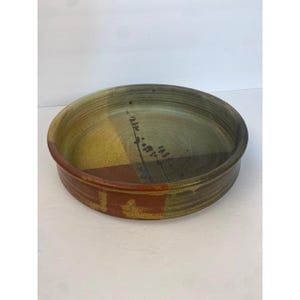 May include: A round ceramic shallow bowl featuring a glazed finish with a color palette of muted olive green, mustard yellow, and deep terracotta red. The interior surface displays a dark linear pattern with small dots, and the exterior rim is smooth.