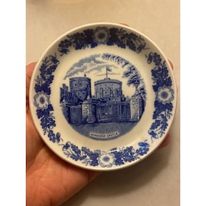 May include: A small, round, white plate with a blue floral border and a central image of Windsor Castle. The castle image is in blue and white, with the words "Windsor Castle" at the bottom. The plate is held in a hand.