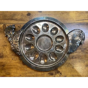 May include: Silver-toned metal egg serving tray with a circular design and ten egg-shaped indentations. The tray features a rooster head and tail design on either side, adding a decorative touch to the serving piece.