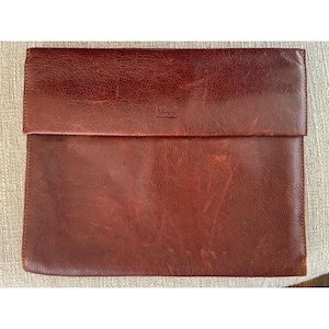 May include: A brown leather document case with a flap closure. The case has a textured surface and a logo embossed on the upper right corner. The leather has a worn appearance, with variations in color and some visible scratches.
