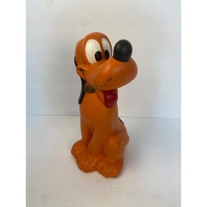 May include: A molded orange dog figurine with large white eyes, black pupils, a black nose, and a red tongue. The character has long black ears and a brown collar around its neck, depicted in a seated position with a smooth, matte finish.