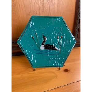 May include: Hexagonal teal plate with a loon illustration. The plate features a black and white loon swimming in a teal pond with reeds. The plate is supported by a gold-colored metal stand.