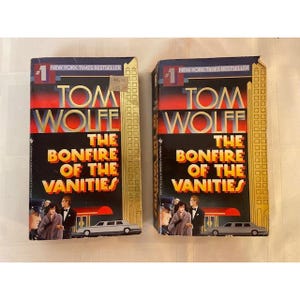 May include: Two identical paperback copies of the book The Bonfire of the Vanities by Tom Wolfe. The covers feature a dark blue and red gradient background, a yellow skyscraper illustration, a white limousine, and a couple in formal evening attire.