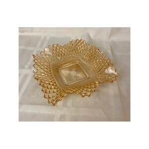 May include: A square amber-colored glass bowl featuring a raised diamond pattern on the exterior sides and a sunburst design radiating from the center of the base. The edges of the bowl are scalloped and pointed, creating a textured appearance.