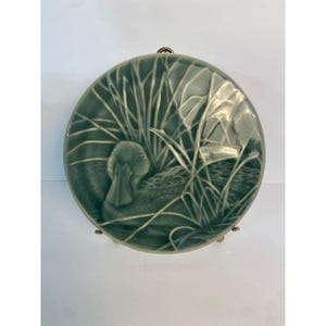 May include: A round ceramic plate featuring a sage green glaze with a relief design of a duck swimming among tall reeds. The surface has a glossy finish with darker green accents highlighting the textured details of the bird and the foliage.