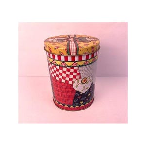 May include: A cylindrical metal tin featuring a colorful patchwork design with red, white, and yellow patterns. The lid displays a central bow graphic, while the body includes checkered, plaid, and heart-shaped motifs with holly leaf accents.