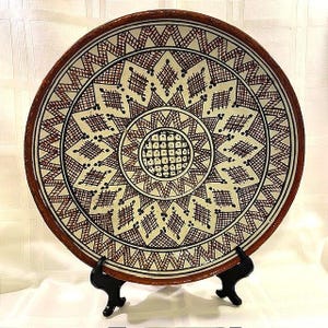 May include: A decorative ceramic plate with a geometric pattern in brown and black on a cream background. The plate has a brown rim and is displayed on a black stand. The design features diamond and star shapes.