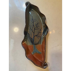 May include: A ceramic leaf-shaped dish with a hand-painted tree design. The dish features a color palette of brown, green, and blue. The rim has a dark brown trim, and the base is a reddish-brown color.