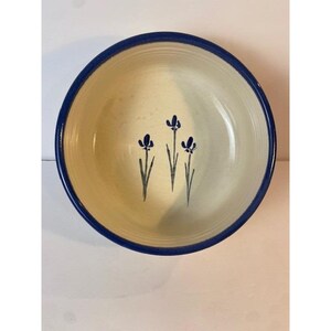 May include: A round ceramic bowl with a cream-colored interior and a dark blue rim. The center of the bowl features a hand-painted design of three blue iris flowers with green stems. The bowl has subtle horizontal ridges along the inner walls.