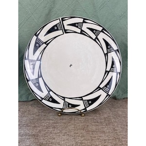 May include: A white ceramic plate with a black geometric design around the rim. The design features repeating black shapes with small circular details. The plate is displayed on a gold-colored metal stand.