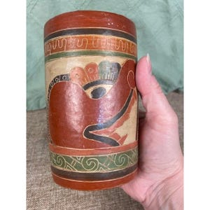 May include: A cylindrical, hand-painted ceramic vase with a reddish-brown exterior. The vase features intricate designs in black, green, and cream colors. The design includes a stylized bird or animal figure.