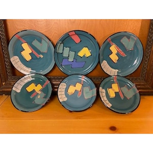 May include: A set of six round ceramic plates featuring a teal background with abstract geometric shapes in yellow, pink, light blue, and purple, accented by a white curved band with thin black lines along the edge of each plate.