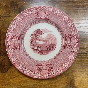 May include: A round, red and white decorative plate with a landscape scene in the center. The plate has a detailed border with floral and architectural elements. The central image depicts a tree, figures, and a castle.