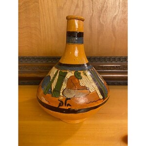 May include: Hand-painted ceramic vase with a bulbous base and a narrow neck. The vase features a warm orange background with a black band and colorful painted figures. The design includes a person, green plants, and other abstract elements. A unique decorative piece.